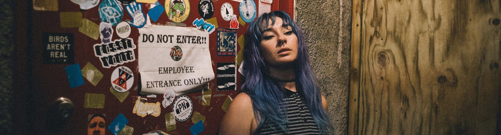 A young person with teal hair standing beside a double door at a punk rock bar covered in band stickers
