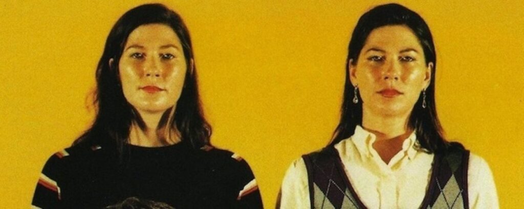 An image of sisters, Kim and Kelley Deal, sitting in front of a vibrant yellow background.