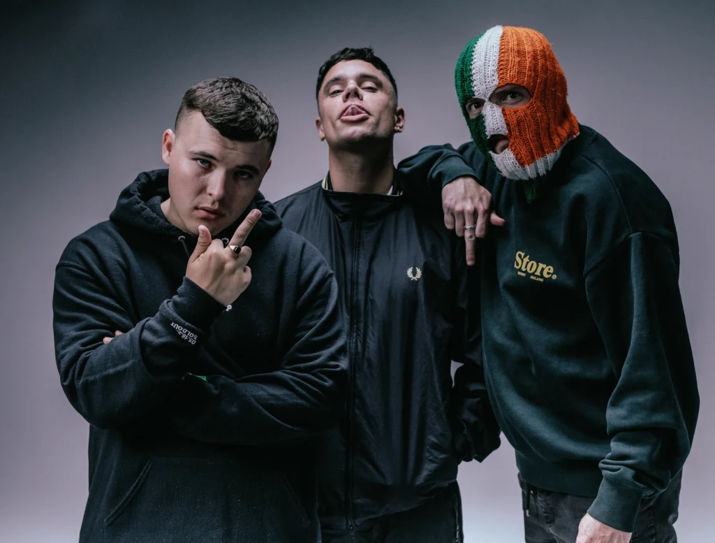 The rap trio, Kneecap, standing in front of a moody gray background.