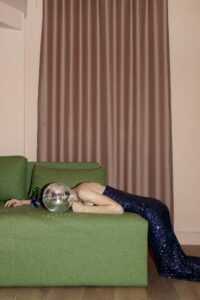 A person in a squint dress laying on a green couch with a disco covering her face.